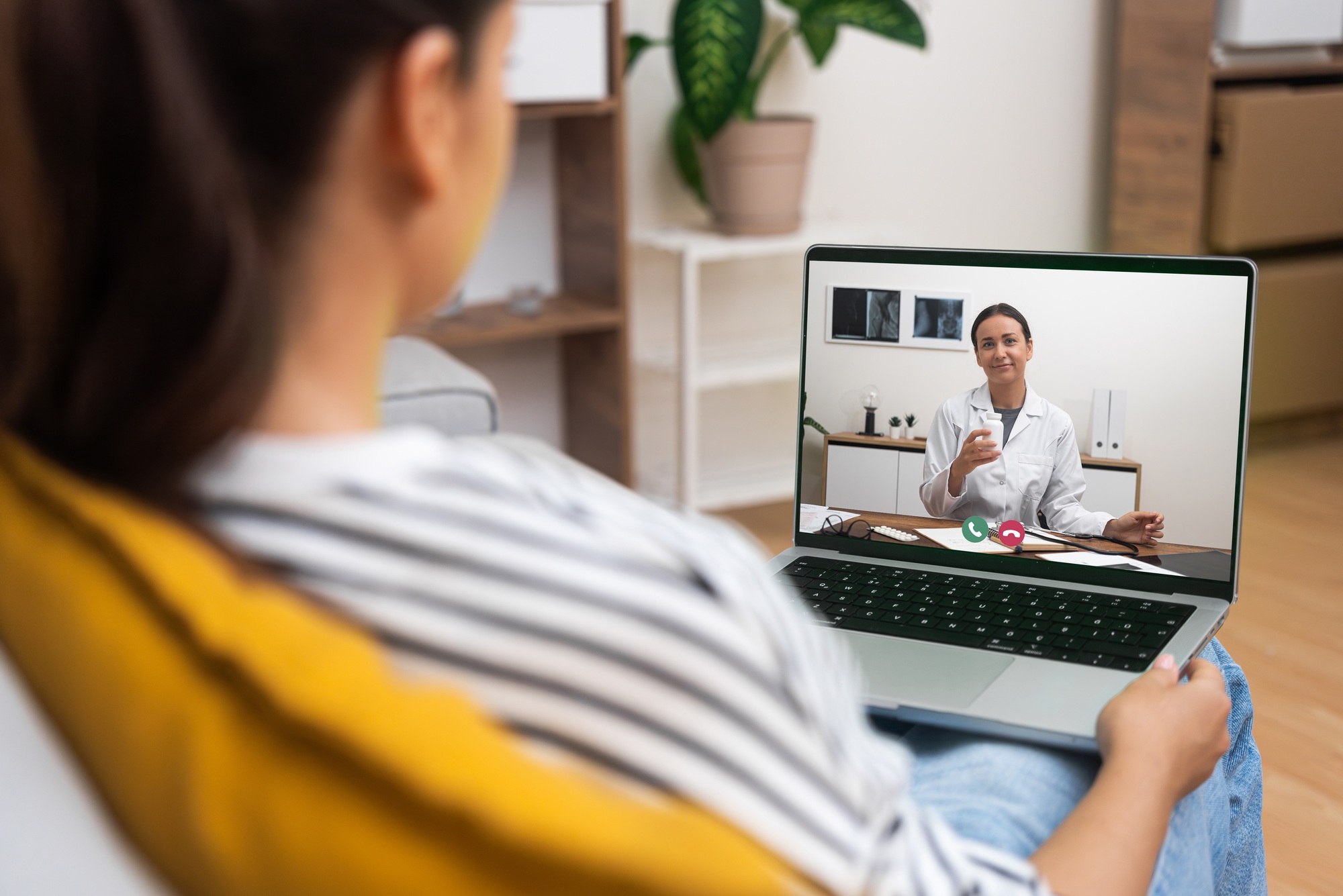 A young woman in a home environment listens to a doctor on a telehealth video call on her laptop.